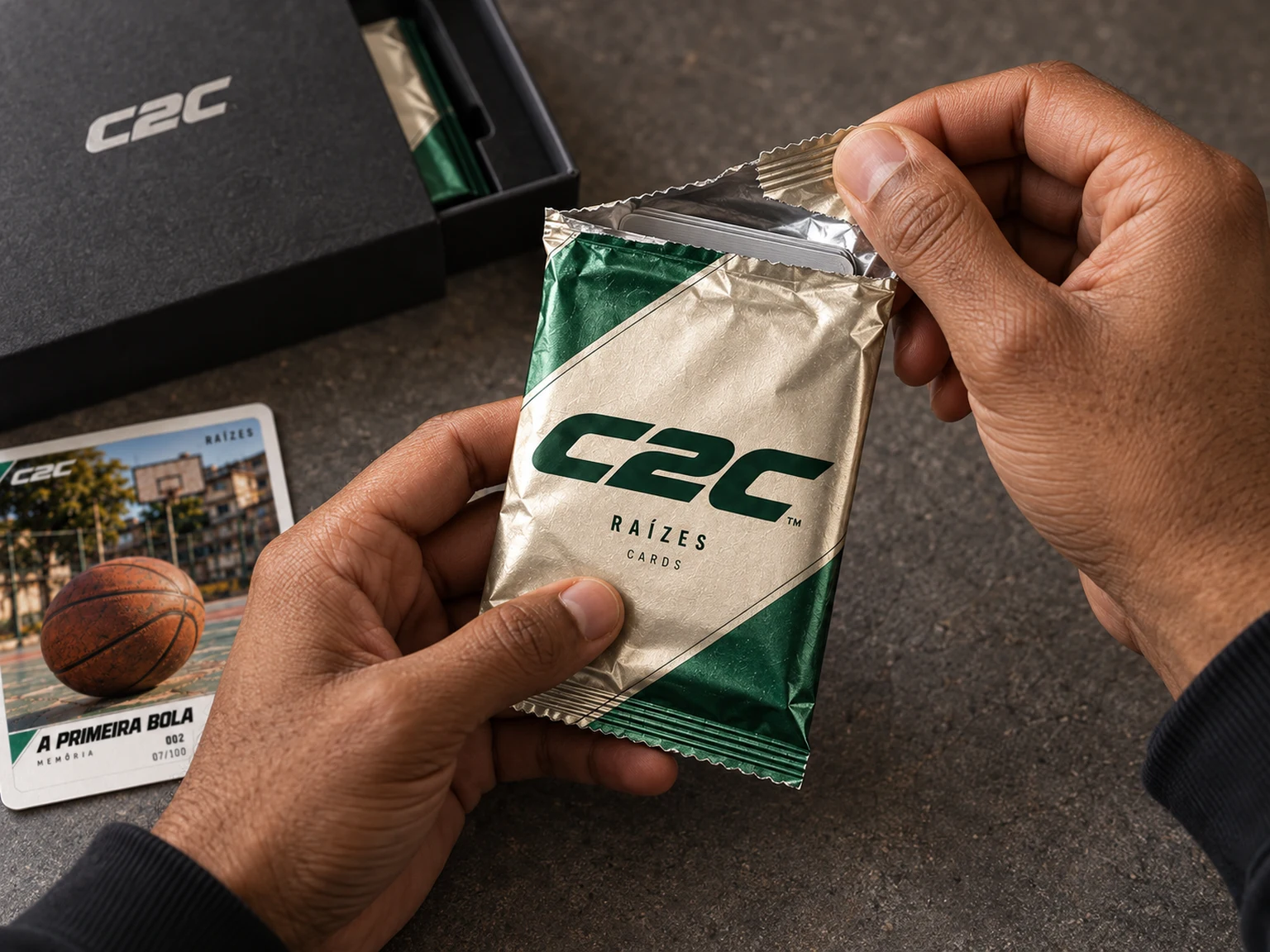 Mãos abrindo um pack metalizado C2C, com caixa preta e card ao fundo
